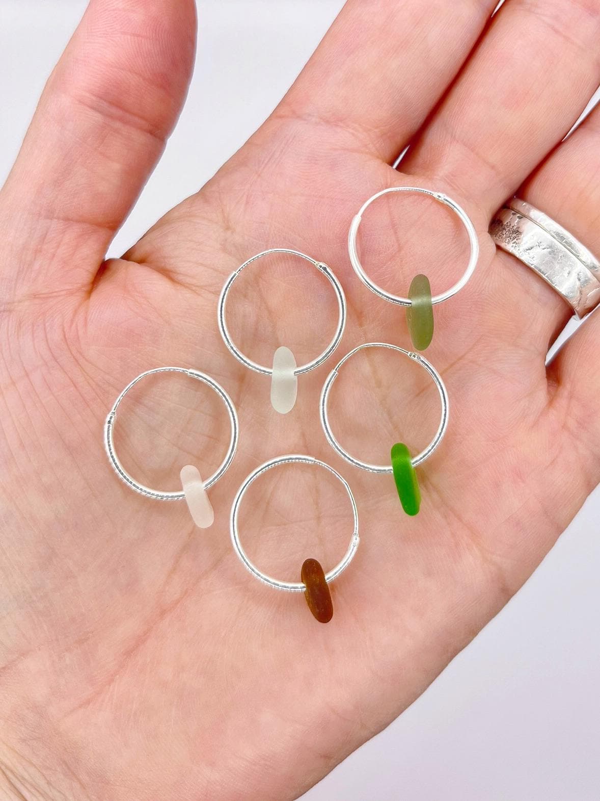 Hand holding five silver hoop earrings with coloured sea glass against a white background