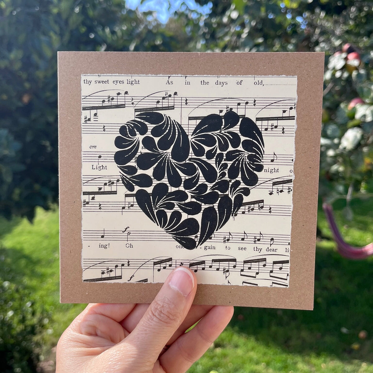 A hand holding a square kraft greetings card with a lino printed heart design in black on music sheet paper mounted on the card. Held against a garden backdrop.