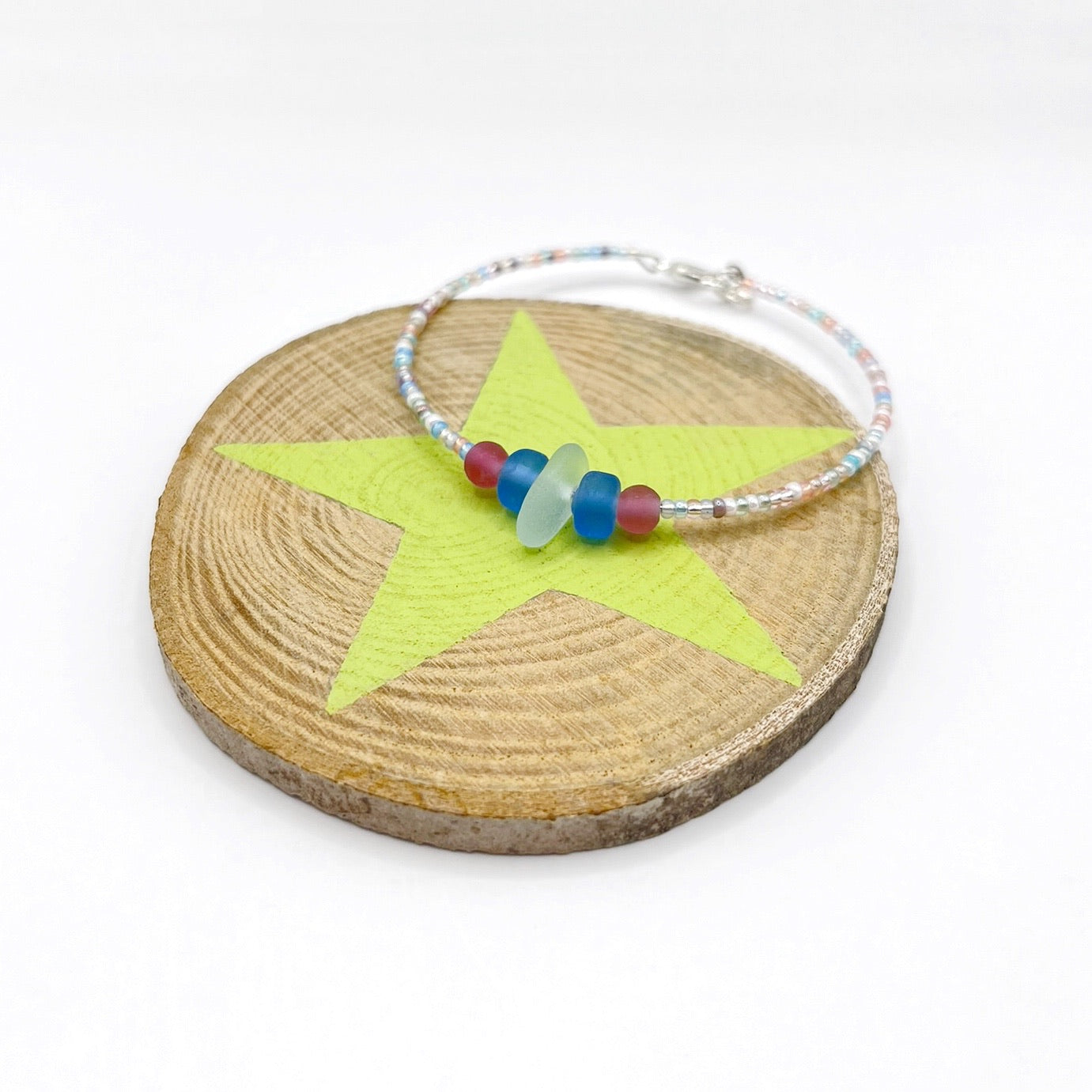 A pastel beaded bracelet with pink and blue frosted glass beads and a piece of sea foam coloured Cornish sea glass, finished with a silver plated lobster clasp and laid on a slice of wood with a pale lime green star design.