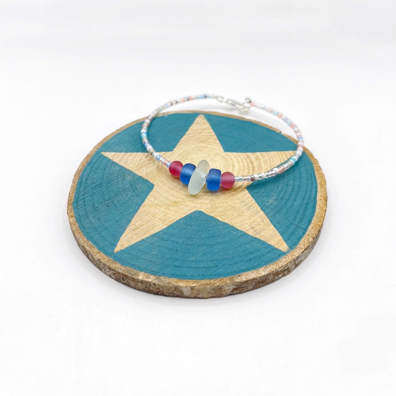 A pastel beaded bracelet with pink and blue frosted glass beads and a piece of sea foam coloured Cornish sea glass, finished with a silver plated lobster clasp and laid on a slice of wood with a blue star design.