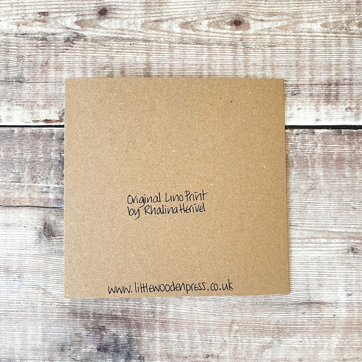 Showing the back of the square brown kraft greetings card with handwritten black text reading 'Original Lino Print by Rhalina Herivel' and 'www.littlewoodenpress.co.uk' Laid on a wooden background.