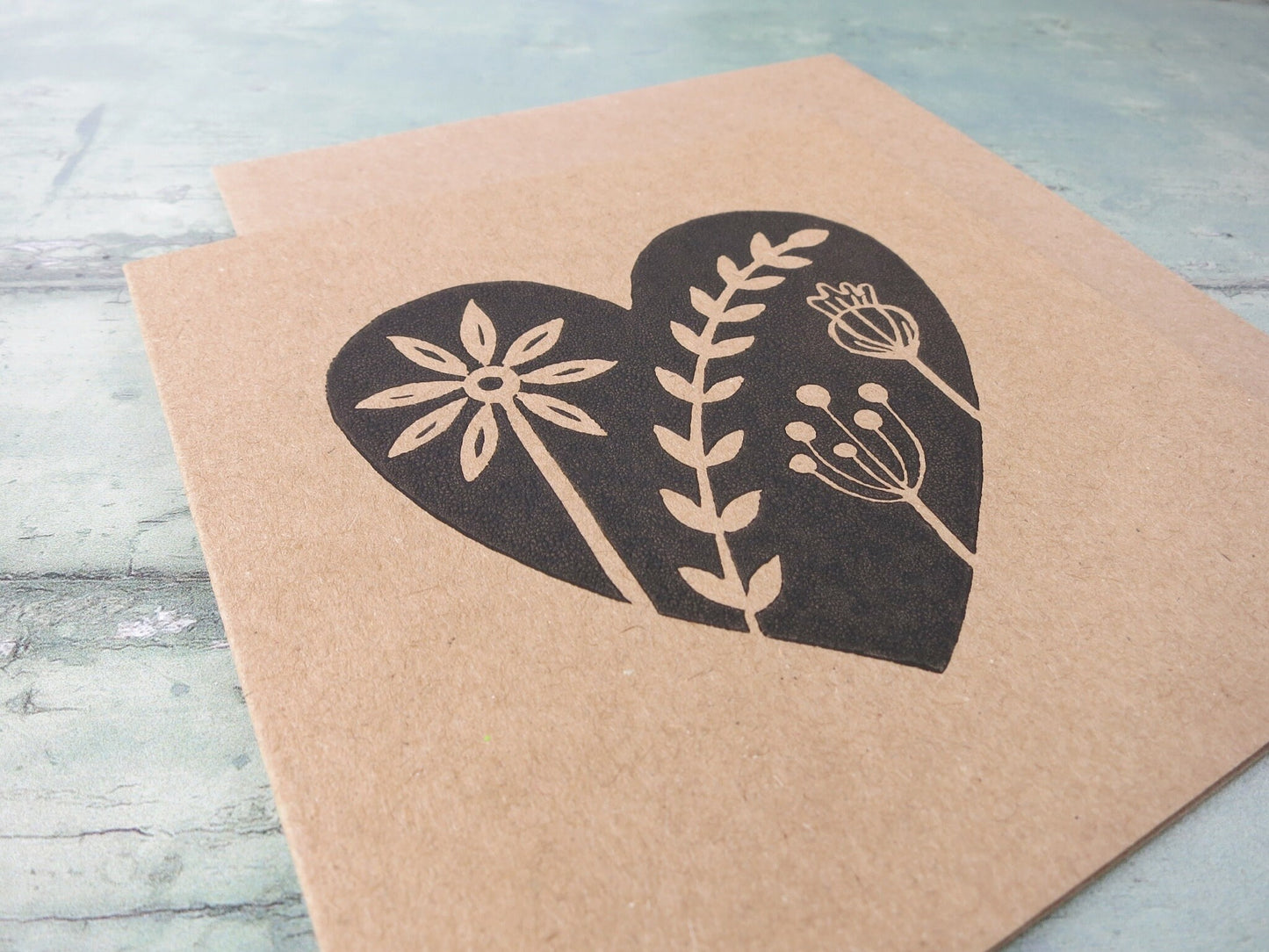 Close up of Kraft paper card with an original black lino printed heart design featuring floral elements laid on a textured surface.