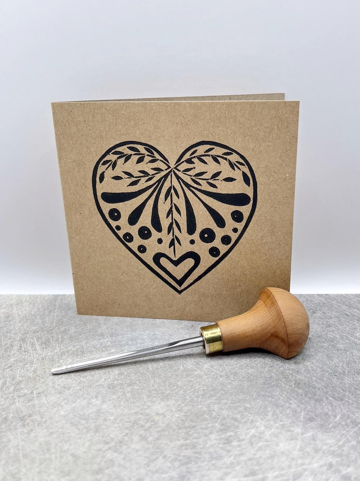 Card with an original lino printed black heart design on Kraft card stood on a textured surface. Photo includes lino cutting tool used.