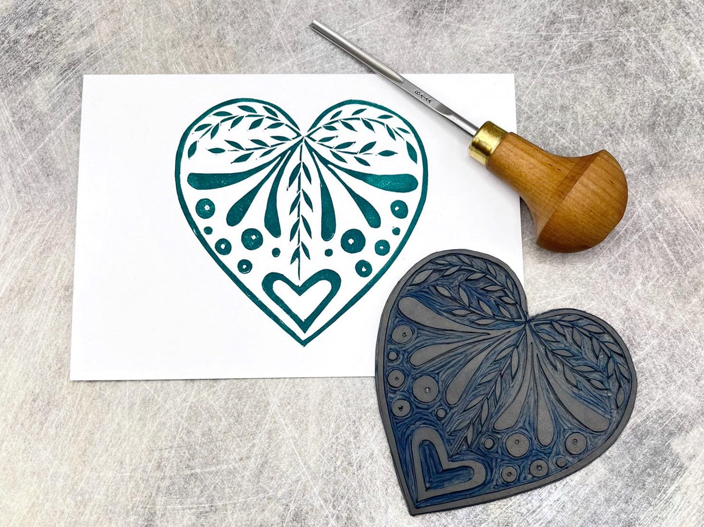 White card with an original lino printed green heart design laid on a textured surface. Photo includes lino and cutting tool.