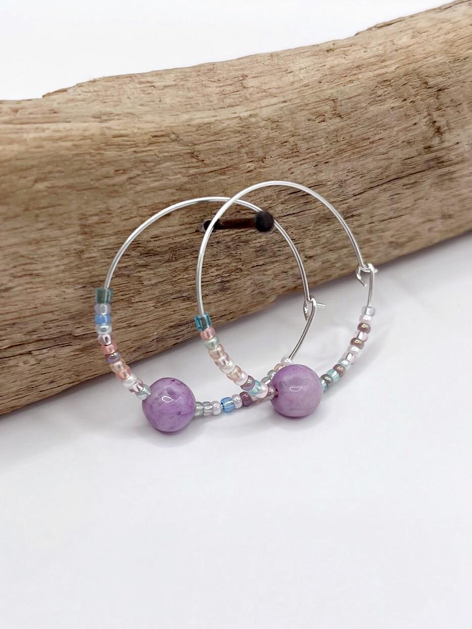 Pair of silver hoop earrings with lilac Jasper and seed beads hanging on driftwood against a light surface.