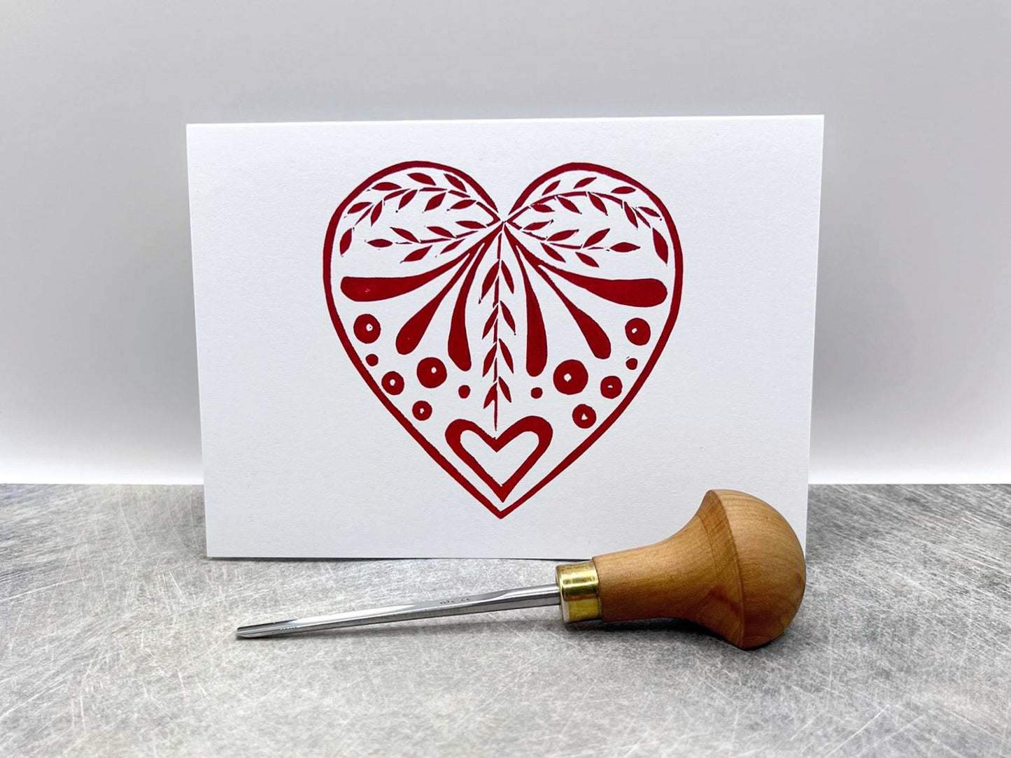 White card with an original lino printed red heart design stood on a textured surface. Photo includes lino cutting tool.