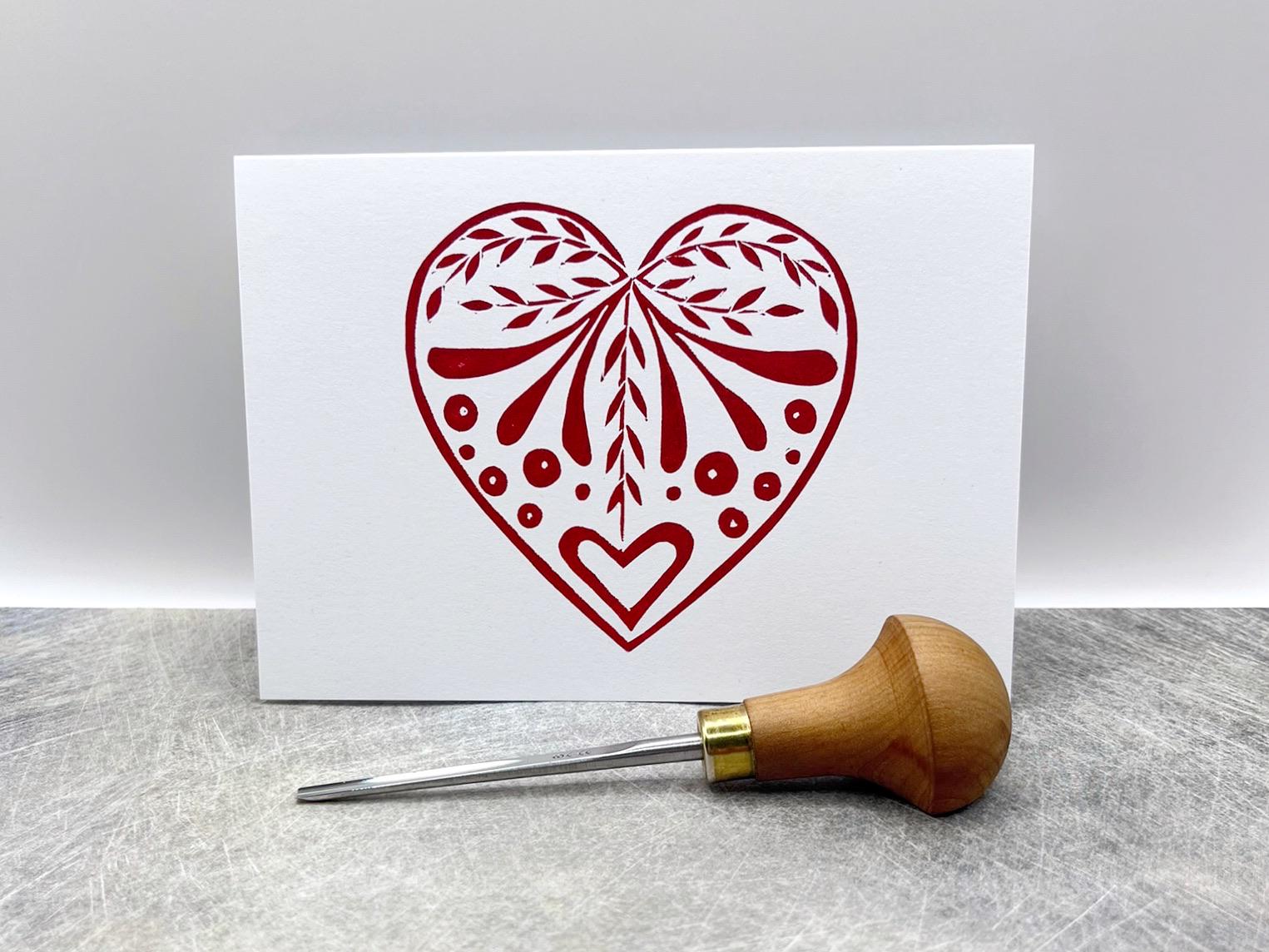 White card with an original lino printed red heart design stood on a textured surface. Photo includes lino cutting tool.