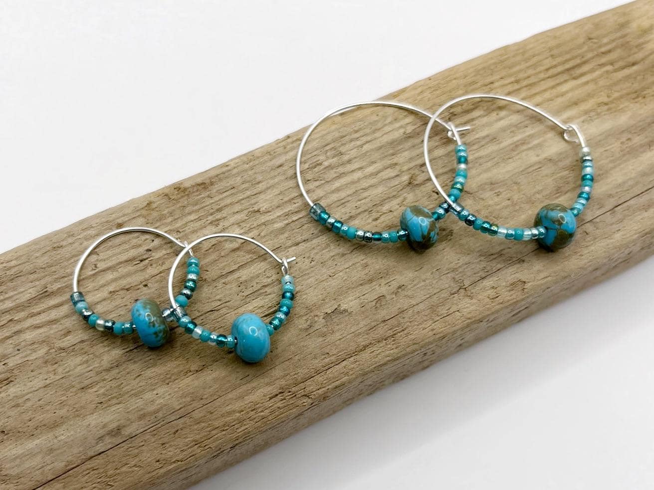 Two pairs of turquoise beaded hoop earrings laid on driftwood