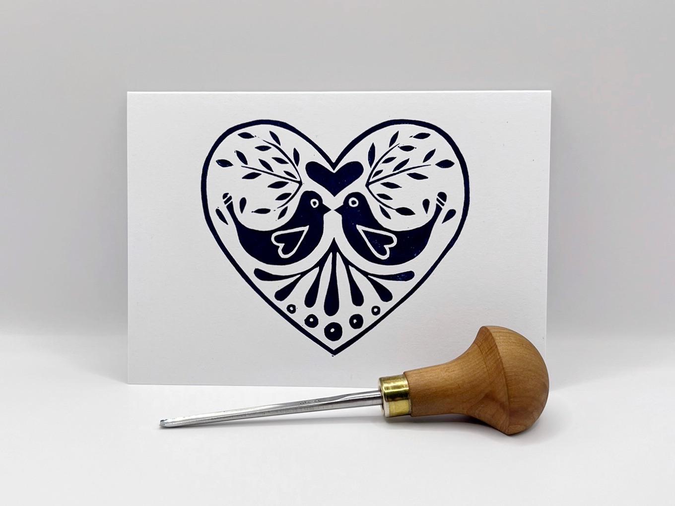 Card with a blue heart design featuring two birds stood against a white background. Photo includes lino cutting tool used.