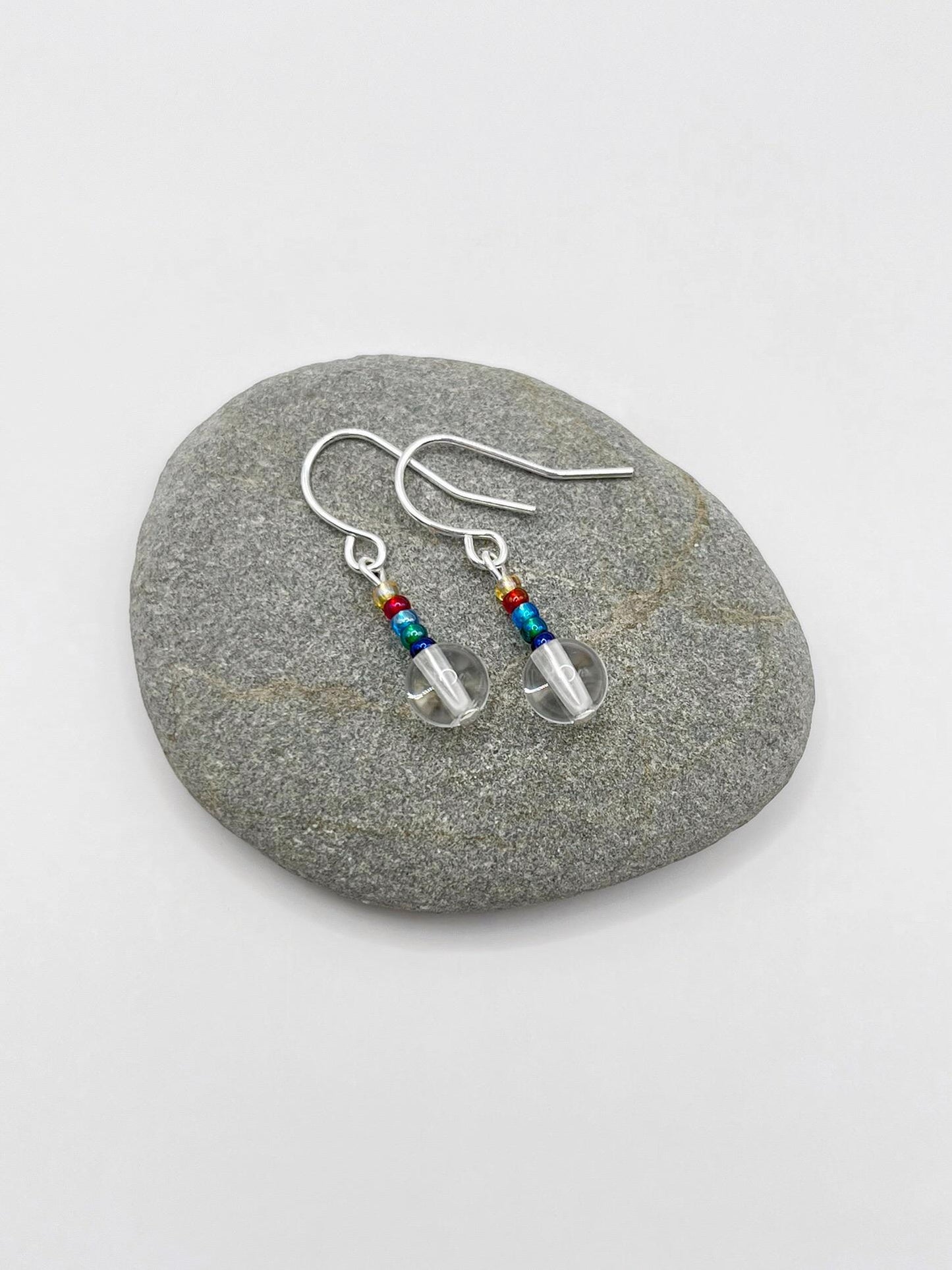 Pair of silver earrings with clear crystal and rainbow seed beads laid on a grey pebble