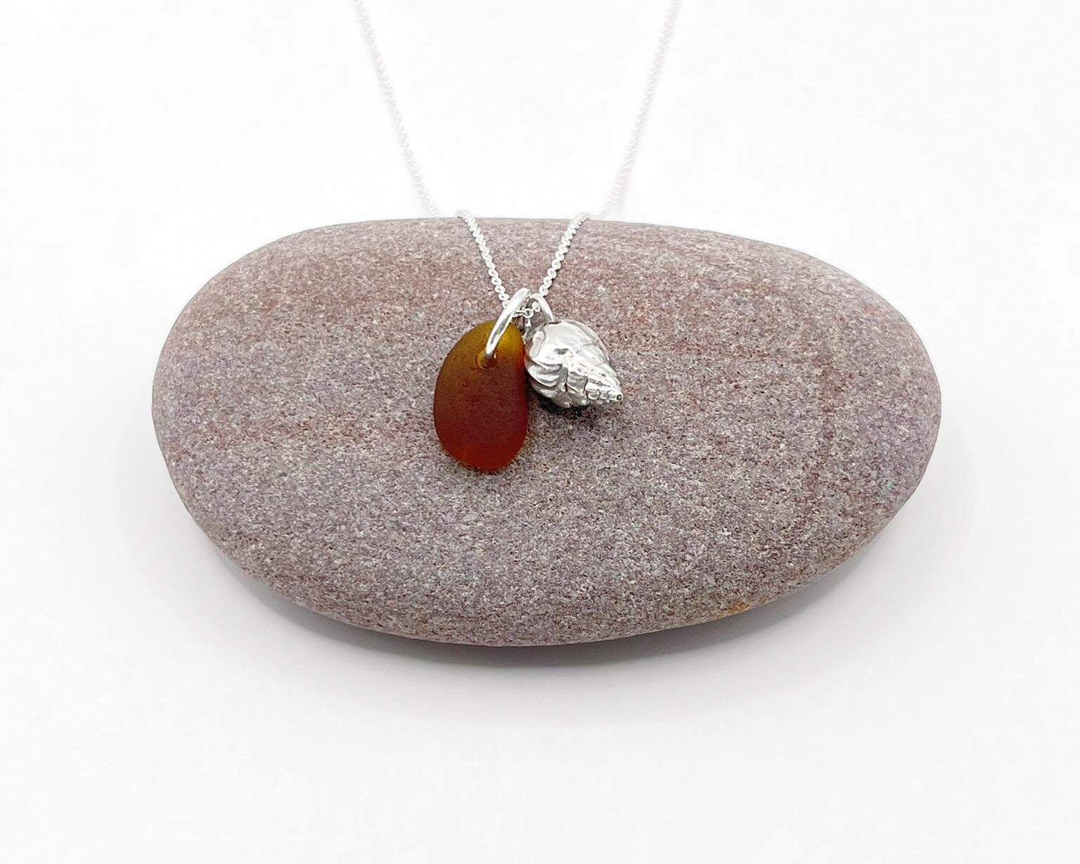 Necklace with burnt orange sea glass and silver shell pendant laid on a purple pebble against a white background