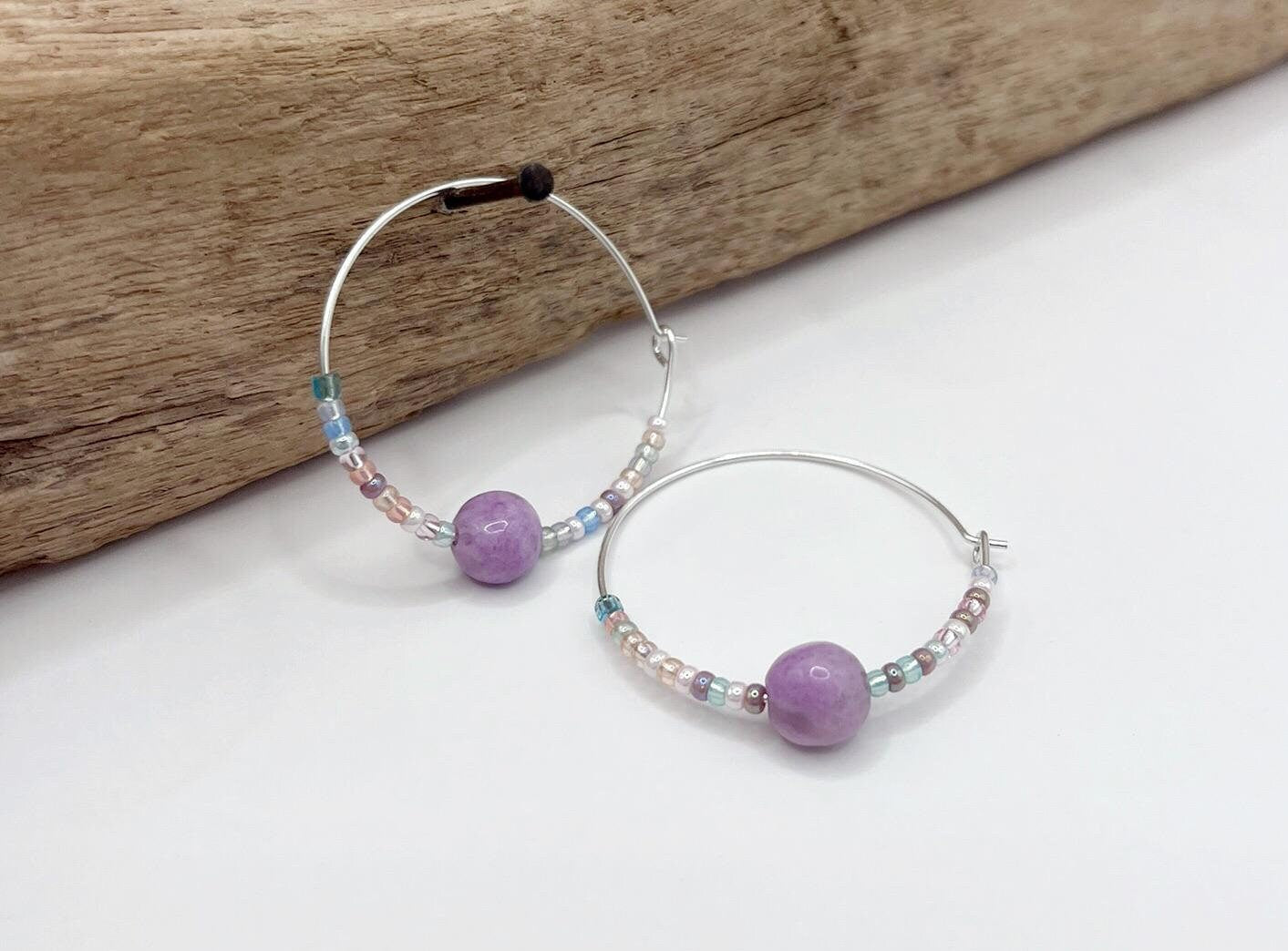 Pair of silver hoop earrings with lilac Jasper and seed beads hanging on driftwood against a light surface.