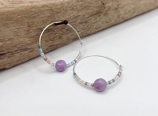 Pair of silver hoop earrings with lilac Jasper and seed beads hanging on driftwood against a light surface.