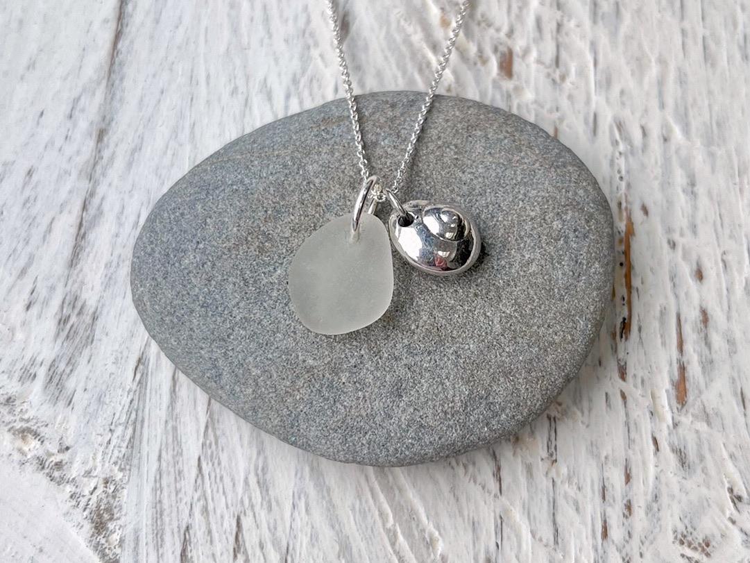 Necklace with a silver shell and sea glass pendant laid on a grey pebble against a textured surface