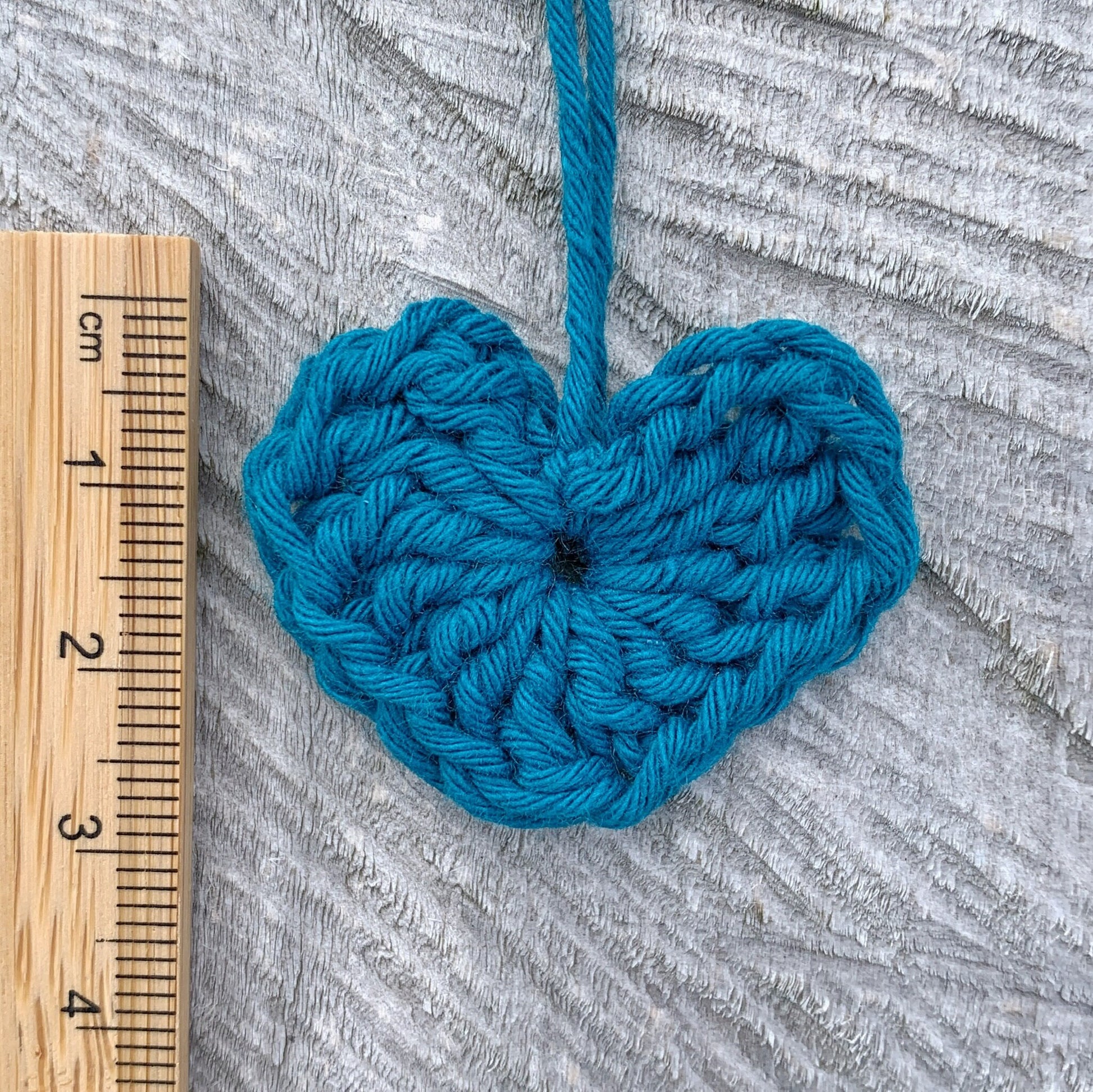 Teal crocheted heart with a wooden ruler for scale on a textured grey background