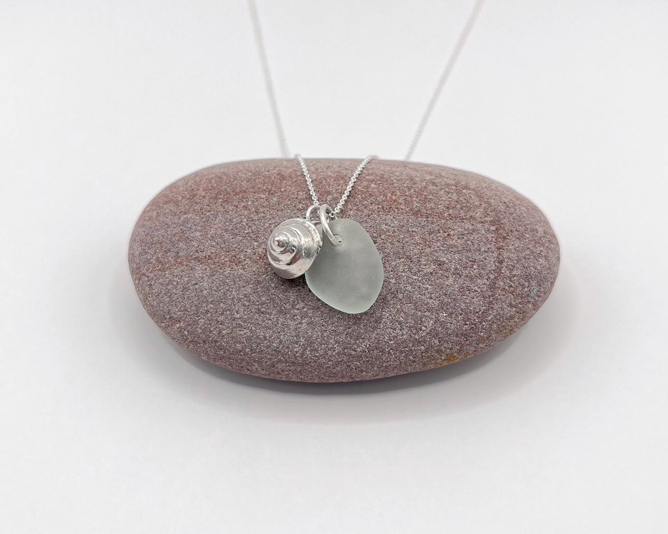 Necklace with a silver shell and seaglass pendant laid on a pebble against a white background