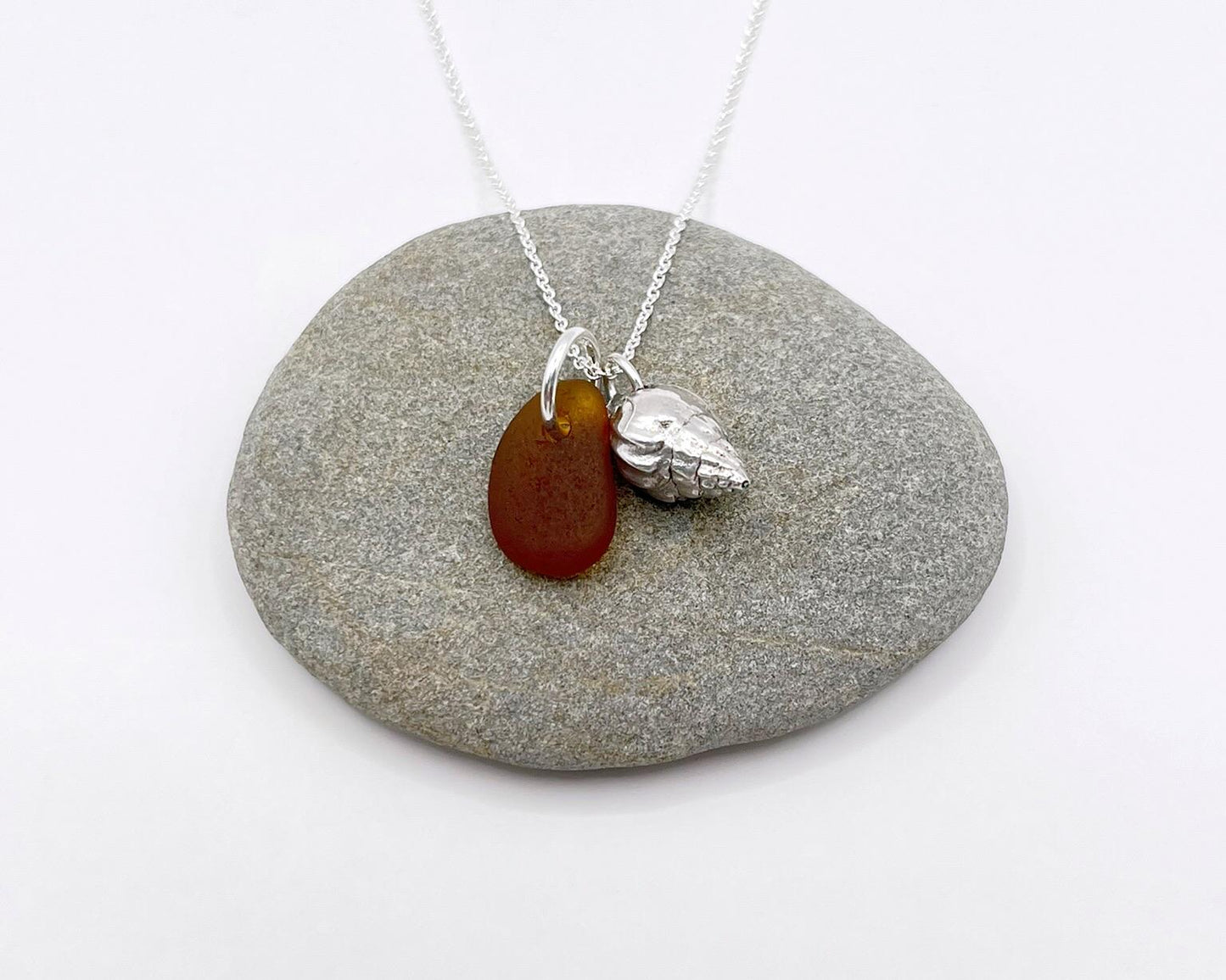 Necklace with burnt orange sea glass and silver shell pendant laid on a grey pebble against a white background