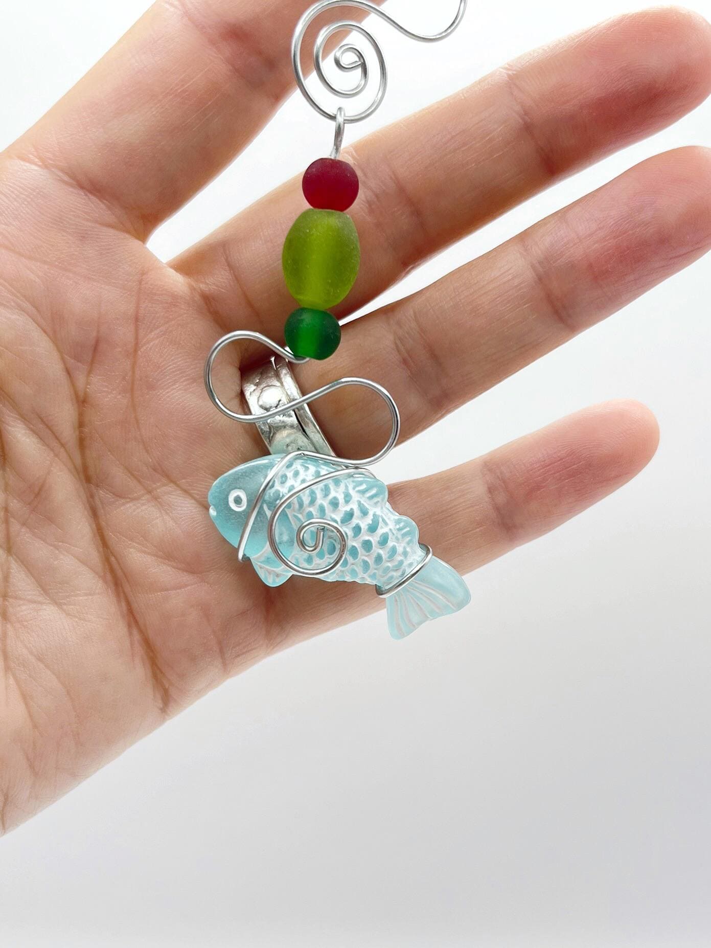 Decorative hanging ornament with a fish design, wire and colourful beads held in a hand against a white background