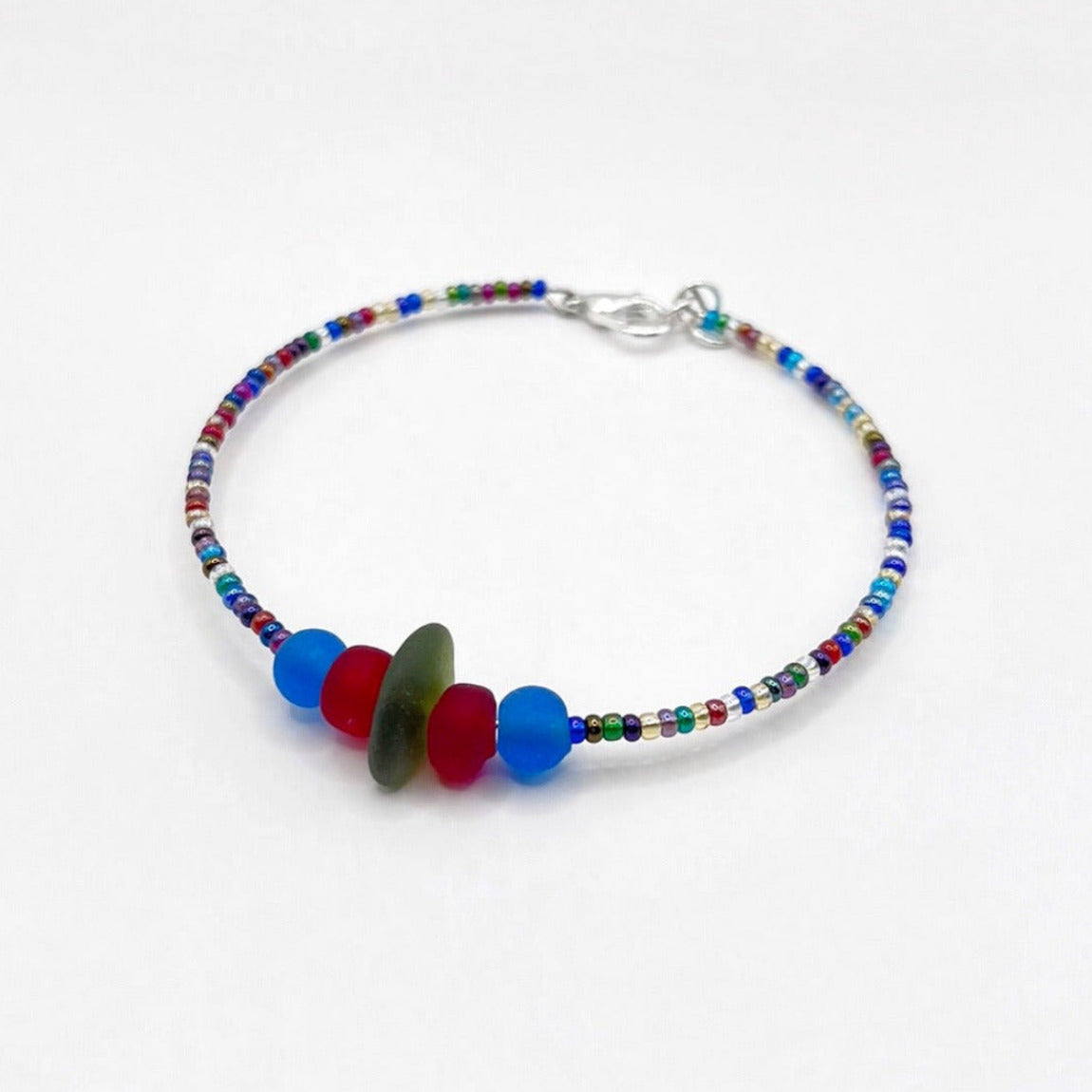 A rainbow coloured beaded bracelet with red and blue frosted glass beads and a piece of olive green coloured Cornish sea glass, finished with a silver plated lobster clasp and laid on a white background.