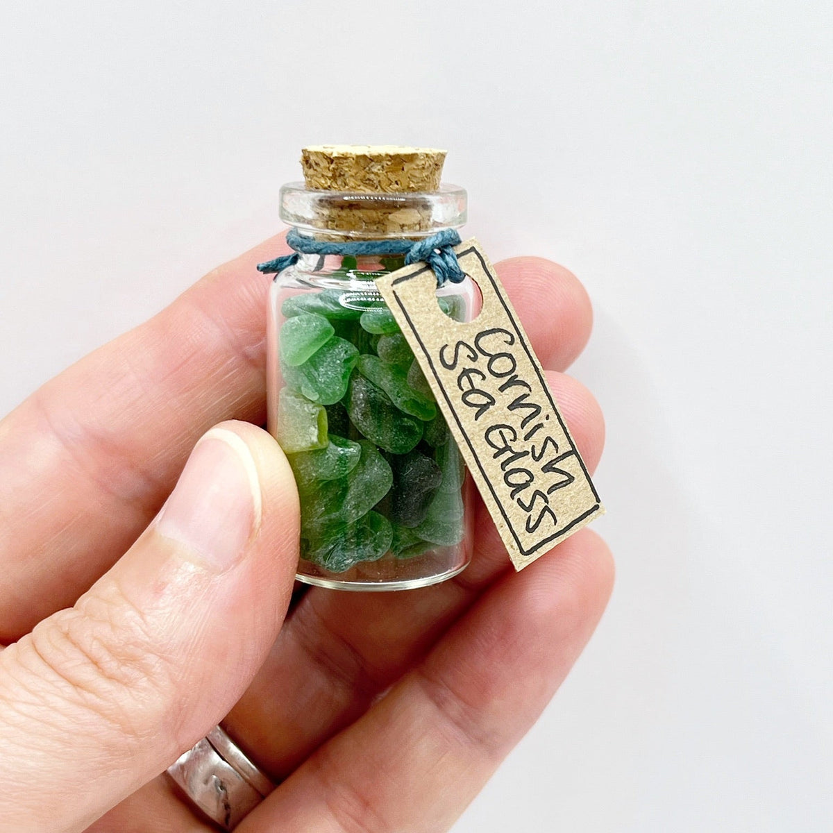 A hand holding a small glass bottle with a cork stopper, filled with green sea glass pieces, and a paper tag attached that reads 'Cornish Sea Glass'.