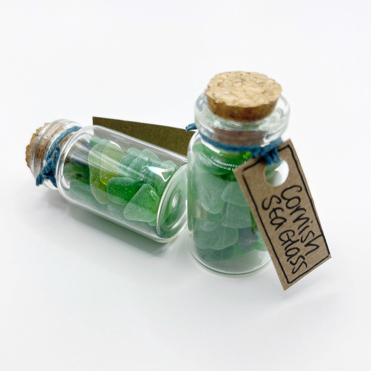 Two small glass bottles with cork stoppers filled with green sea glass, tied with blue string and one has a hanging tag that reads 'Cornish Sea Glass' on a white background.