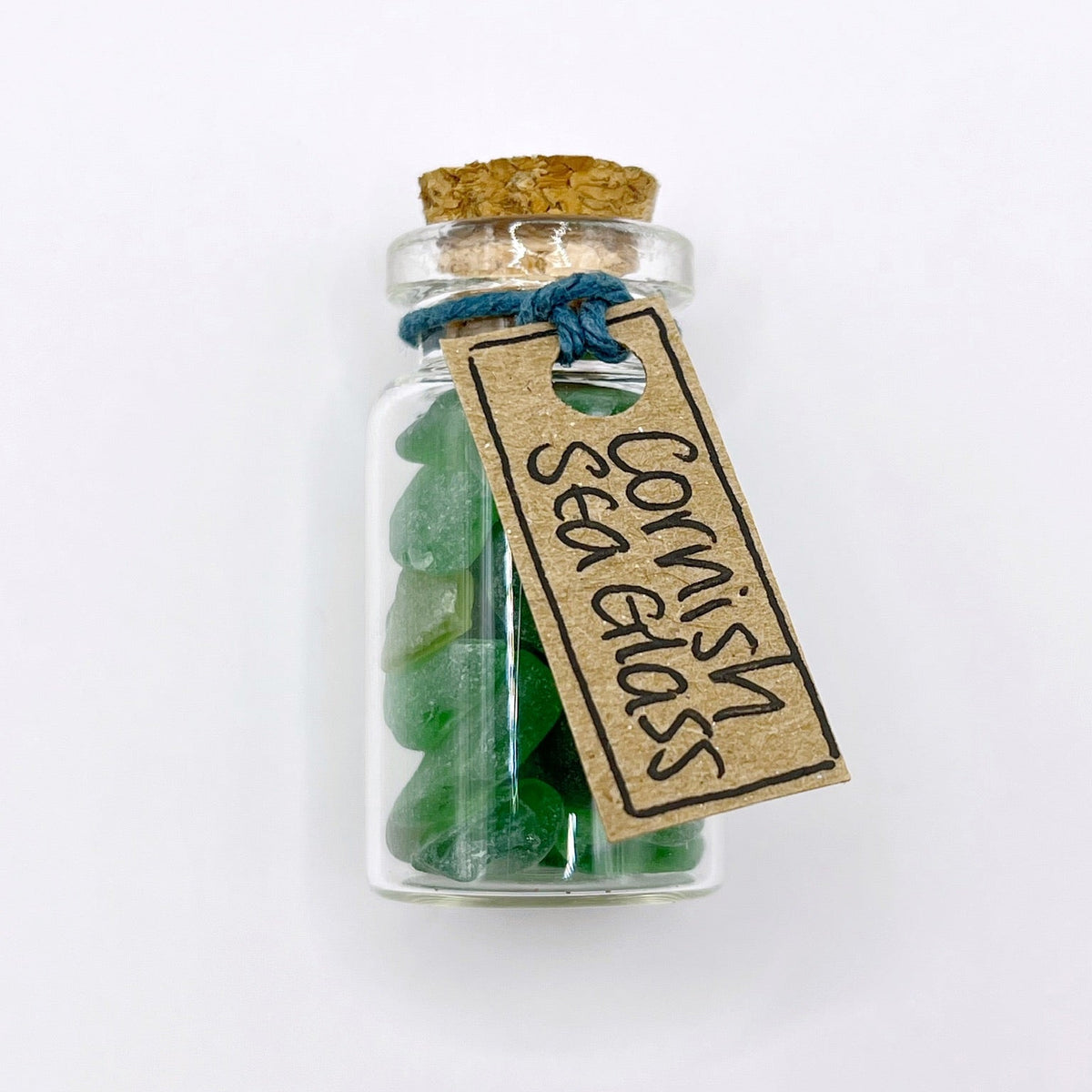 A small clear glass bottle with a cork stopper, filled with green sea glass pieces, tied with blue string and has a kraft paper tag that reads 'Cornish Glass Sea Glass'. The bottle is photographed on a clean white background.