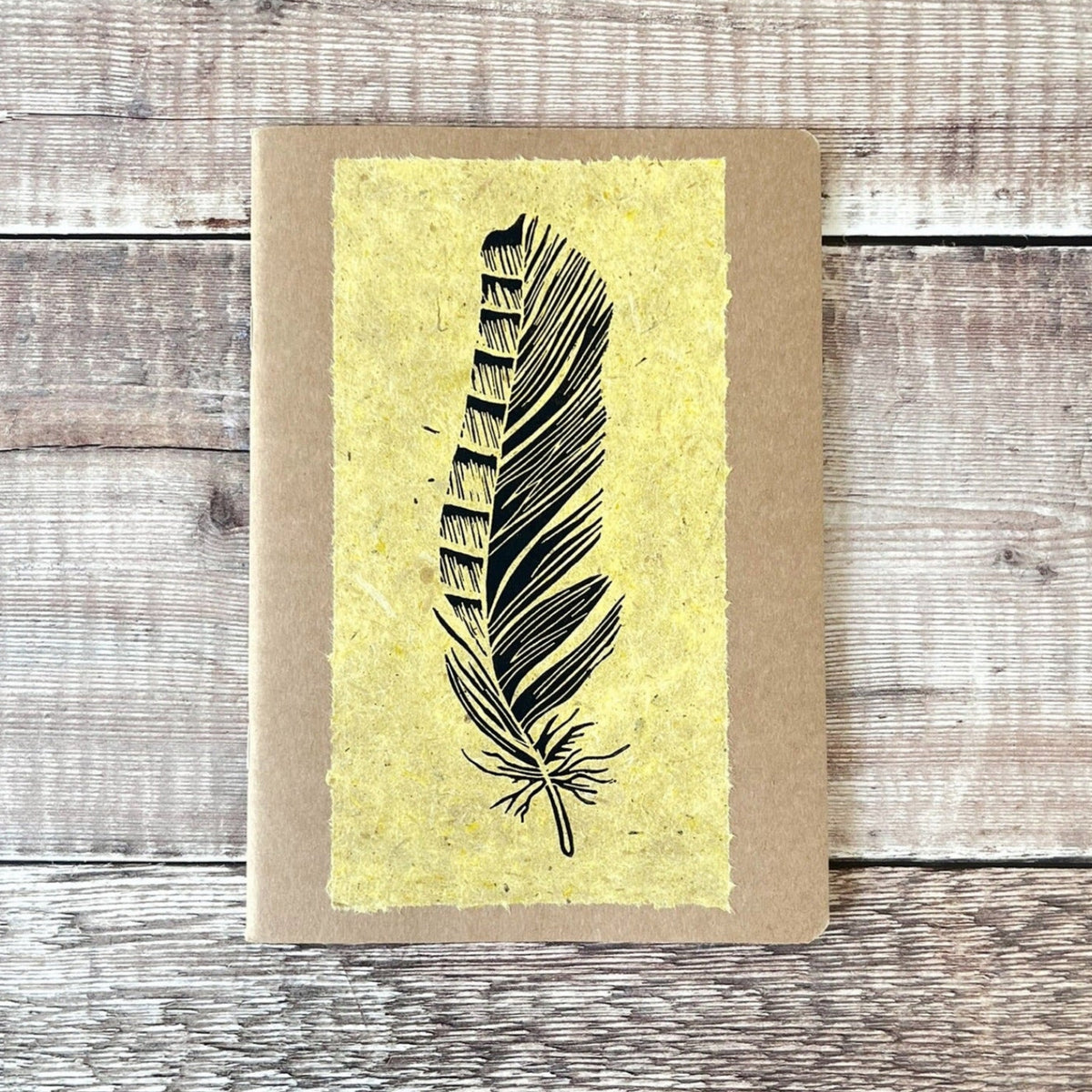 A5 kraft paper notebook or journal with a textured yellow handmade paper panel featuring a black lino ink feather illustration, photographed flat on a rustic wood plank surface.