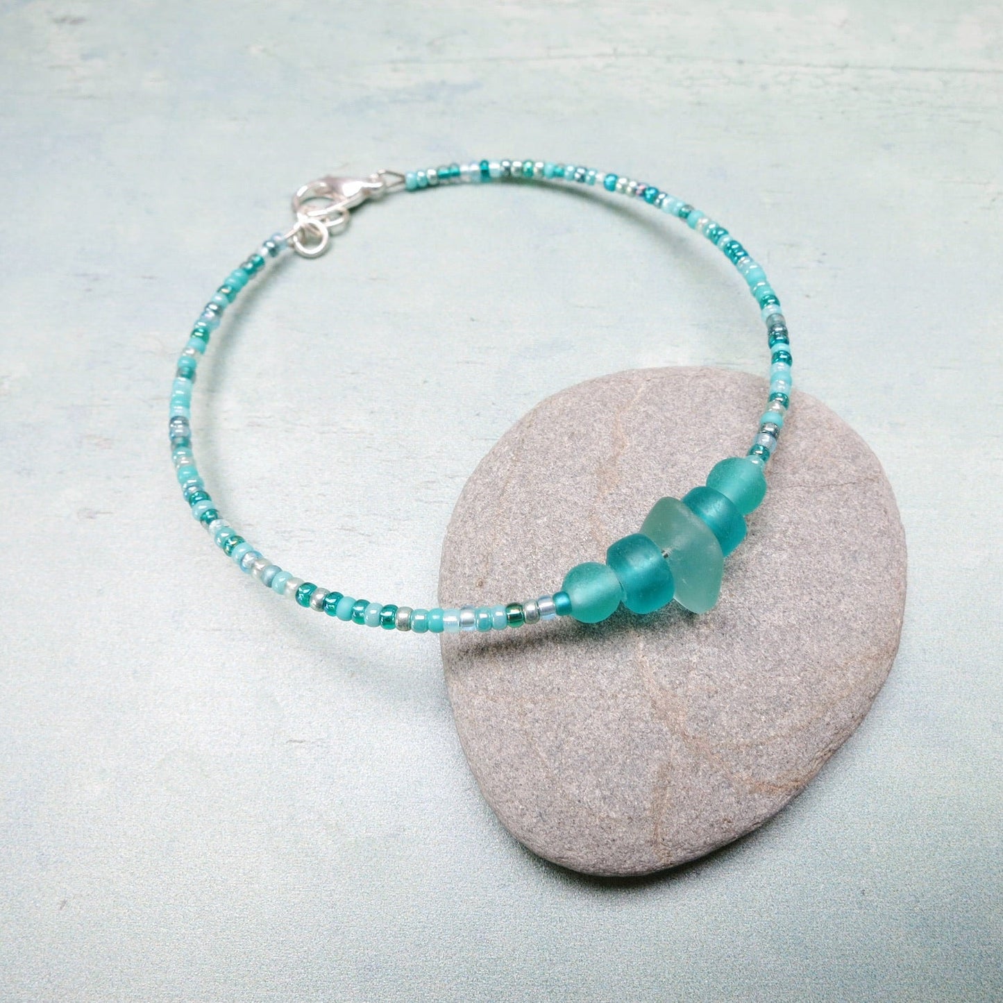 Bracelet with pale sea foam sea glass and sea green and turquoise glass beads laid on a grey pebble against a pale blue background