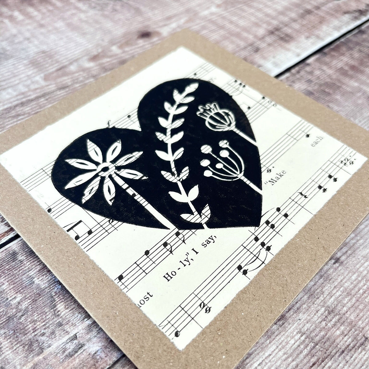Square kraft greeting card with a black floral heart lino print on vintage music paper mounted on the front. Laid on a wooden background.