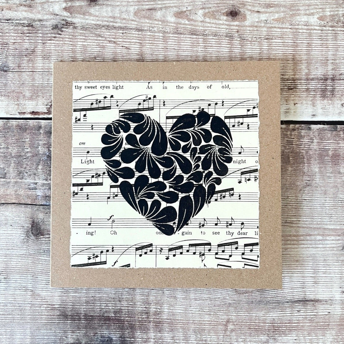 Square kraft greetings card with a lino printed heart design in black on music sheet paper mounted on the card. Laid on a wooden background.