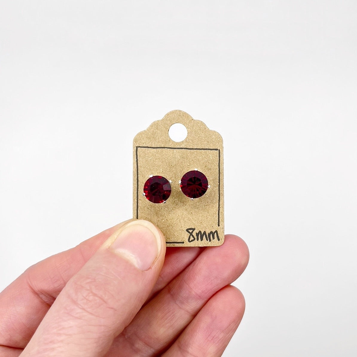 Pair of red cut glass stud earrings on a small cardboard tag held by a hand against a white background