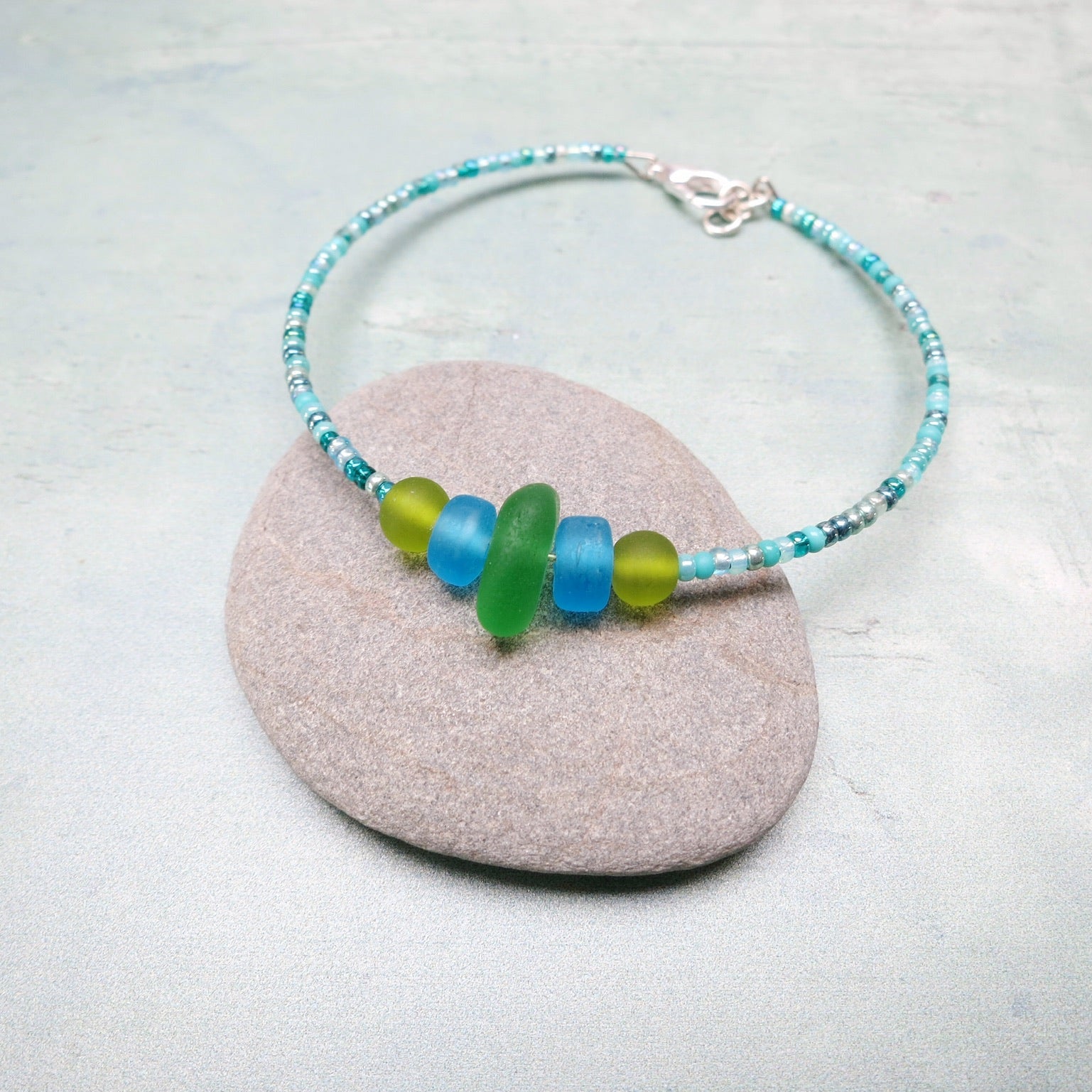 Bracelet with green sea glass and turquoise and green glass beads laid on a purple pebble against a pale blue background