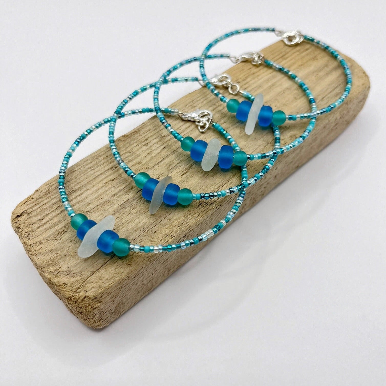 Four bracelets with white sea glass and blue and turquoise beads laid on driftwood against a white background