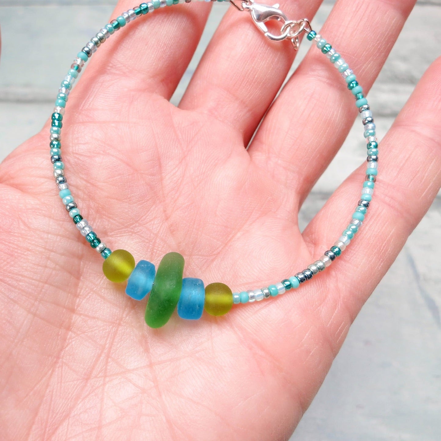 Bracelet with green sea glass and turquoise and green glass beads held in a hand against a pale blue background