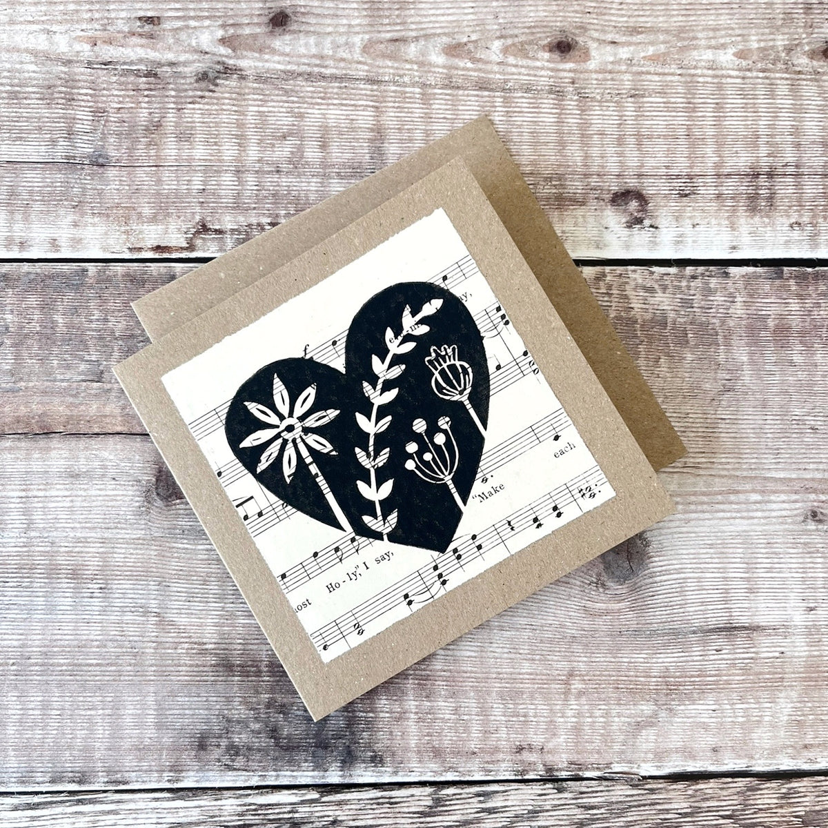 Square kraft greeting card with a black floral heart lino print on vintage music paper mounted on the front. Laid on a matching envelope on a wooden background.