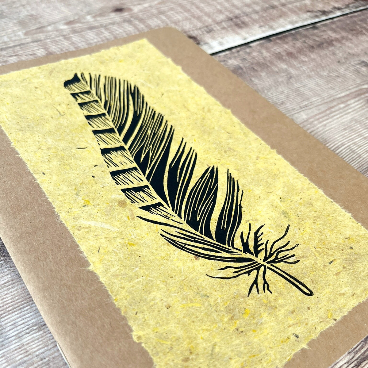 Close up of A5 kraft paper notebook or journal with a textured yellow handmade paper panel featuring a black lino ink feather illustration, photographed flat on a rustic wood plank surface.