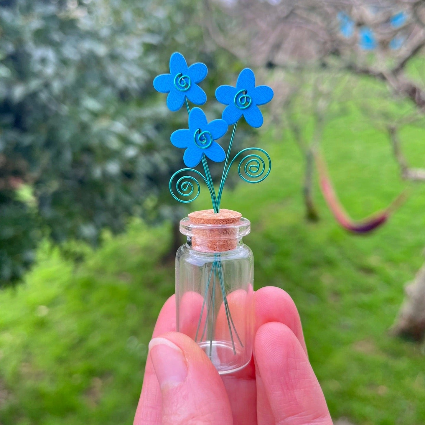 Little glass bottle with three blue wooden flowers held in a hand against a garden background