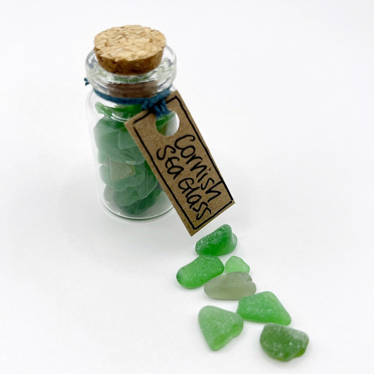 A small clear glass bottle with a cork stopper and a hanging tag reading 'Cornish Sea Glass,' filled with green sea glass, with several loose green sea glass pieces scattered on a white background.