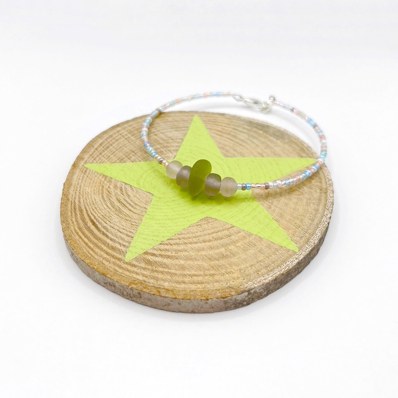 A pastel beaded bracelet with purple and lilac frosted glass beads and a piece of soft lime coloured Cornish sea glass, finished with a silver plated lobster clasp and laid on a slice of wood with a pale lime star design.