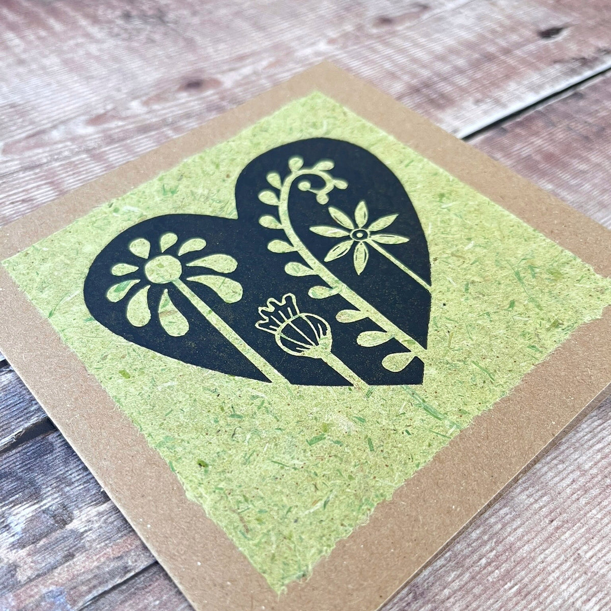 Close up of a square kraft greeting card with a black floral heart lino print on pale green handmade paper mounted on the front. Laid on a wooden background.
