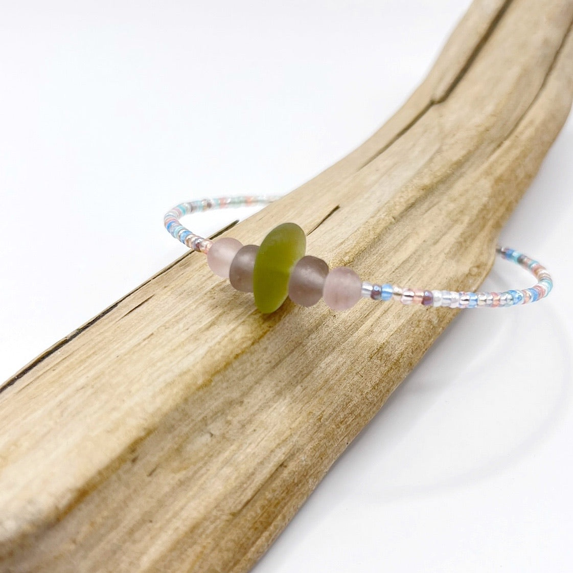 A pastel beaded bracelet with purple and lilac frosted glass beads and a piece of soft lime coloured Cornish sea glass, finished with a silver plated lobster clasp and laid on a piece of driftwood.