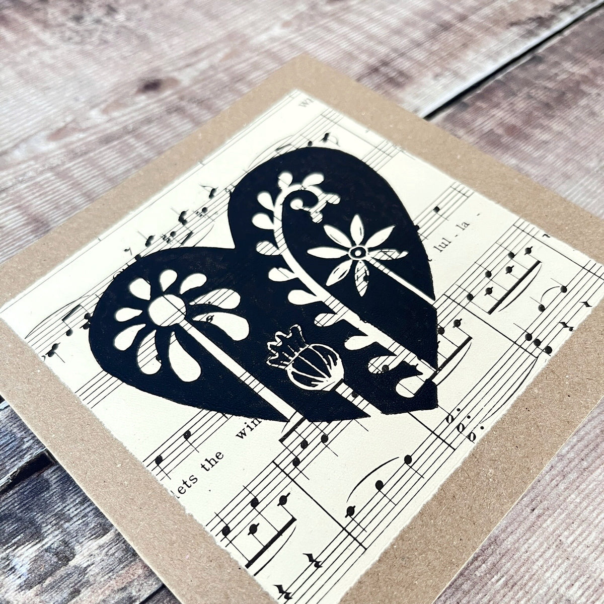 Close up of square kraft greeting card with a black floral heart lino print on vintage music paper mounted on the front. Laid on a wooden background.