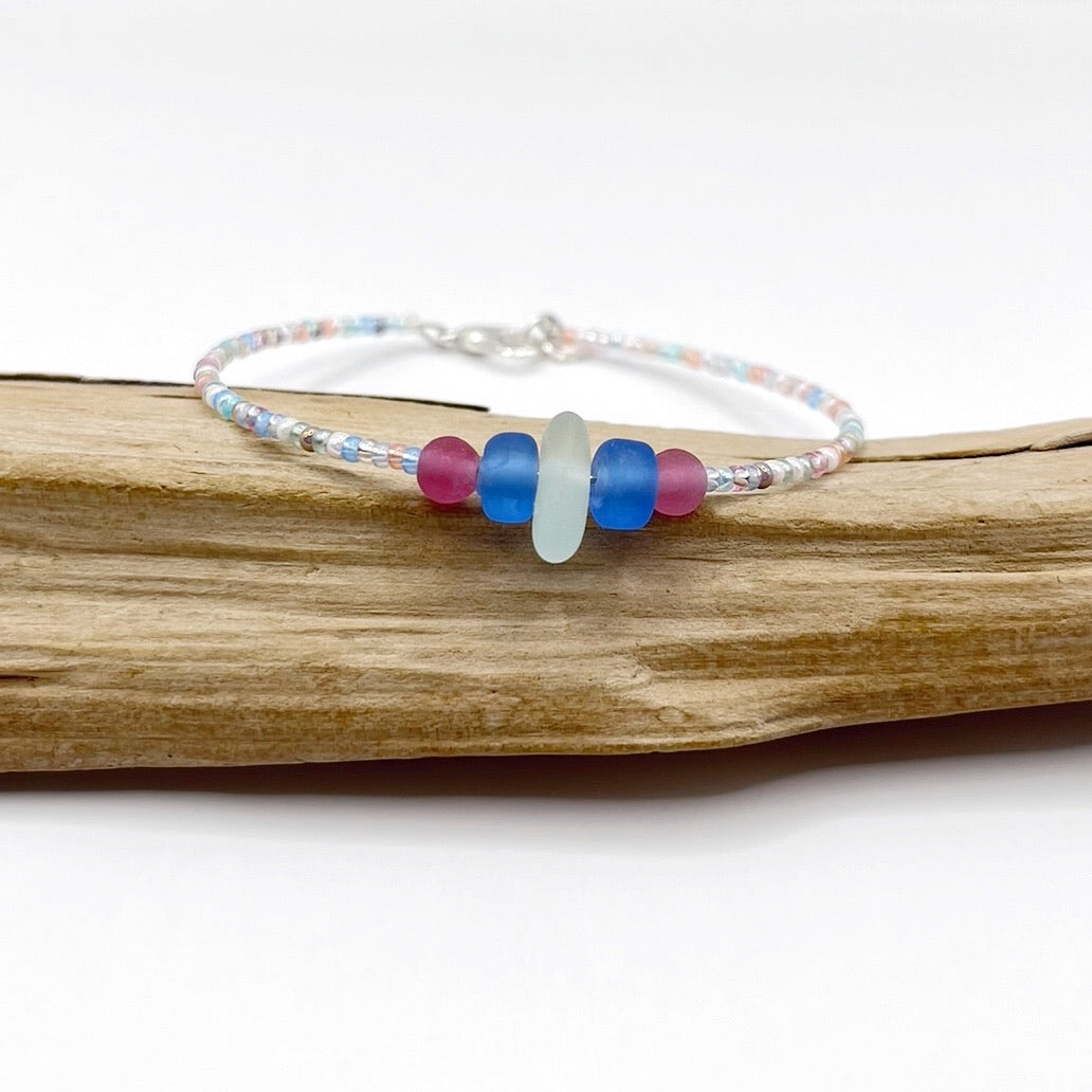 A pastel beaded bracelet with pink and blue frosted glass beads and a piece of sea foam coloured Cornish sea glass, finished with a silver plated lobster clasp and laid on a piece of driftwood.