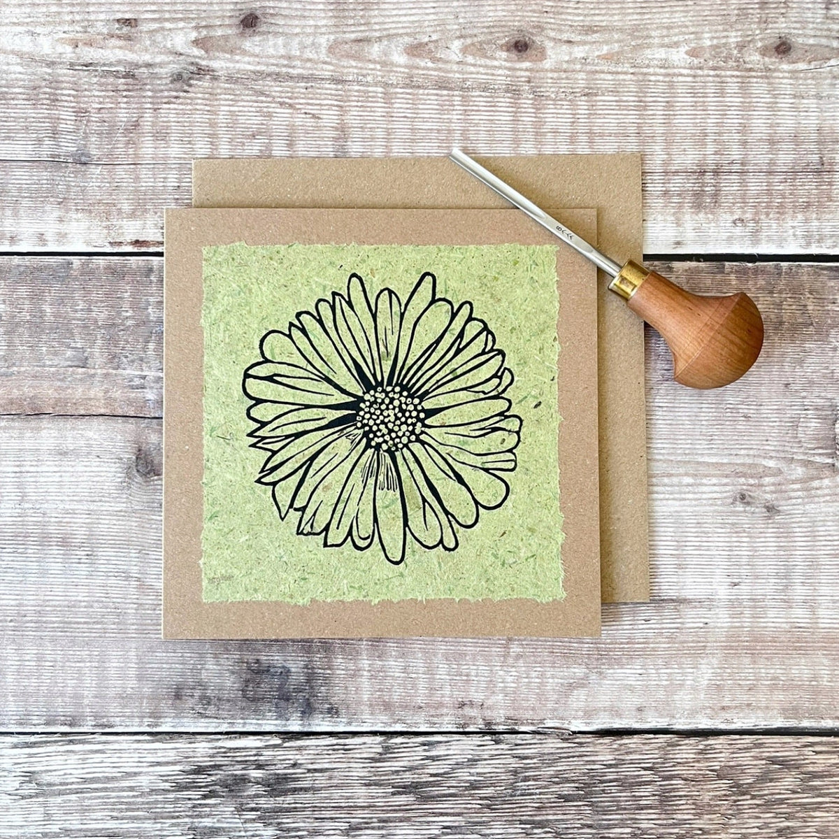 A square handmade art card featuring a black ink lino print of a daisy flower on a textured light green handmade paper, mounted on a brown kraft greetings card, laid flat on a rustic wooden surface. Photo includes the lino cutting tool used.