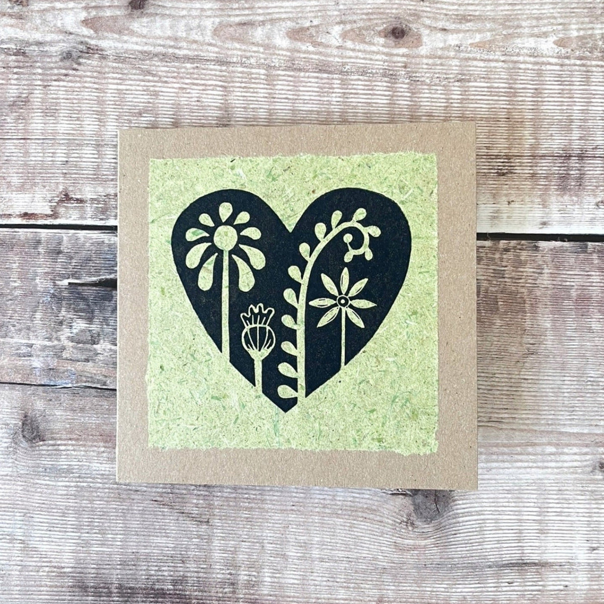 Square kraft greeting card with a black floral heart lino print on pale green handmade paper mounted on the front. Laid on a wooden background.