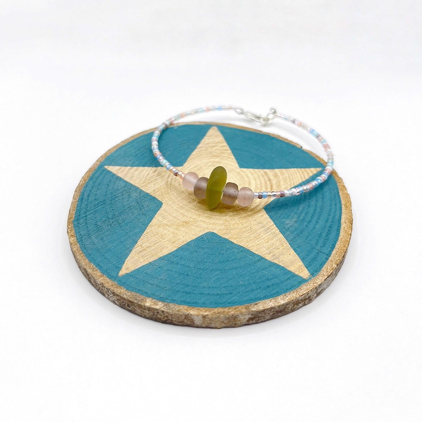 A pastel beaded bracelet with purple and lilac frosted glass beads and a piece of soft lime coloured Cornish sea glass, finished with a silver plated lobster clasp and laid on a slice of wood with a blue star design.