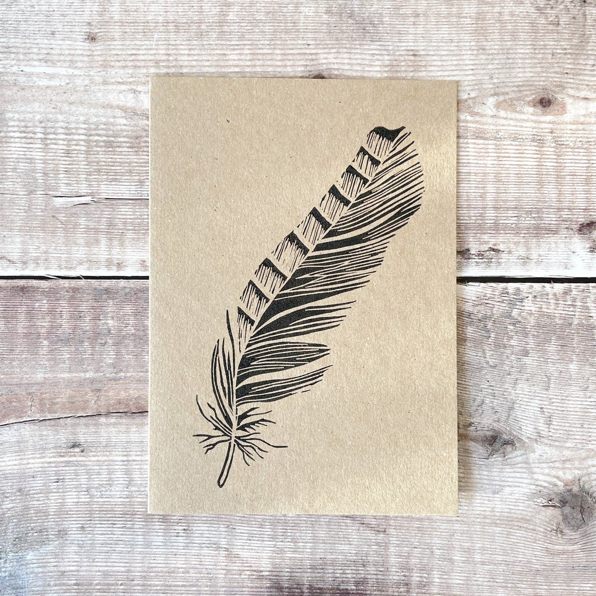 Feather lino print in black printed on a kraft card laid on a wooden surface.