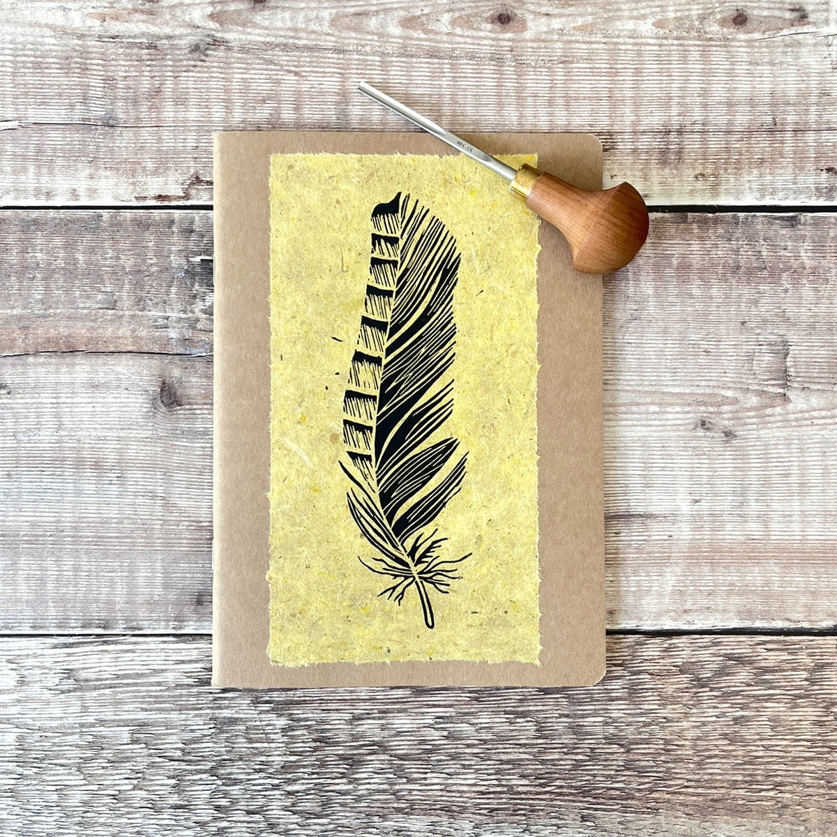 A5 kraft paper notebook or journal with a textured yellow handmade paper panel featuring a black lino ink feather illustration, photographed flat on a rustic wood plank surface. Photo includes the lino cutting tool used.