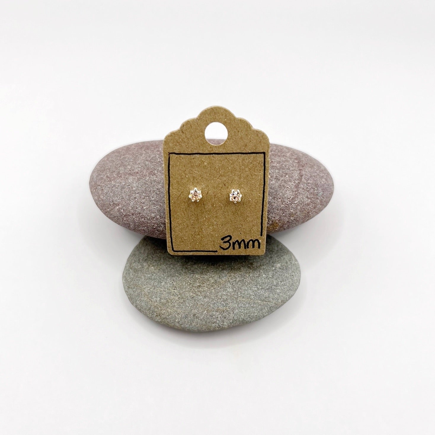 Pair of silver stud earrings on a small cardboard stand with '3mm' text, placed on two stones.
