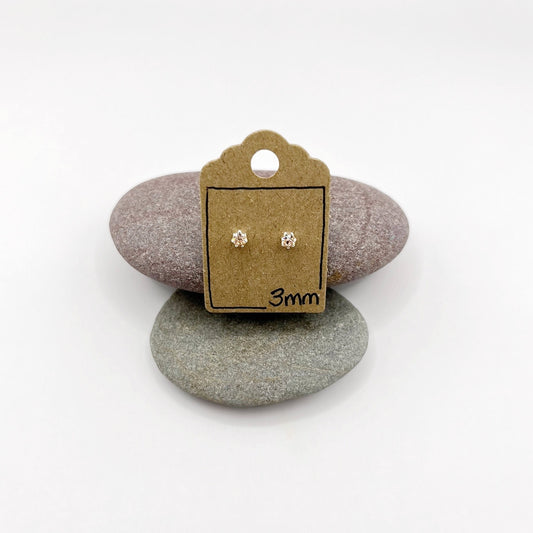 Pair of silver stud earrings on a small cardboard stand with '3mm' text, placed on two stones.